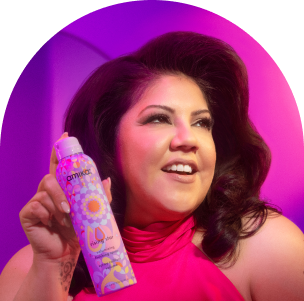 image of model with voluminous, styled hair smiles while holding a can of Amika's "Plus Size" volumizing finishing spray. The model is dressed in a vibrant pink top, and the background features a purple and pink gradient. The "Plus Size" can is prominently displayed in the model's hand, showcasing its colorful, decorative packaging with intricate patterns. The overall image emphasizes the product's volumizing capabilities and the playful, energetic branding of Amika.