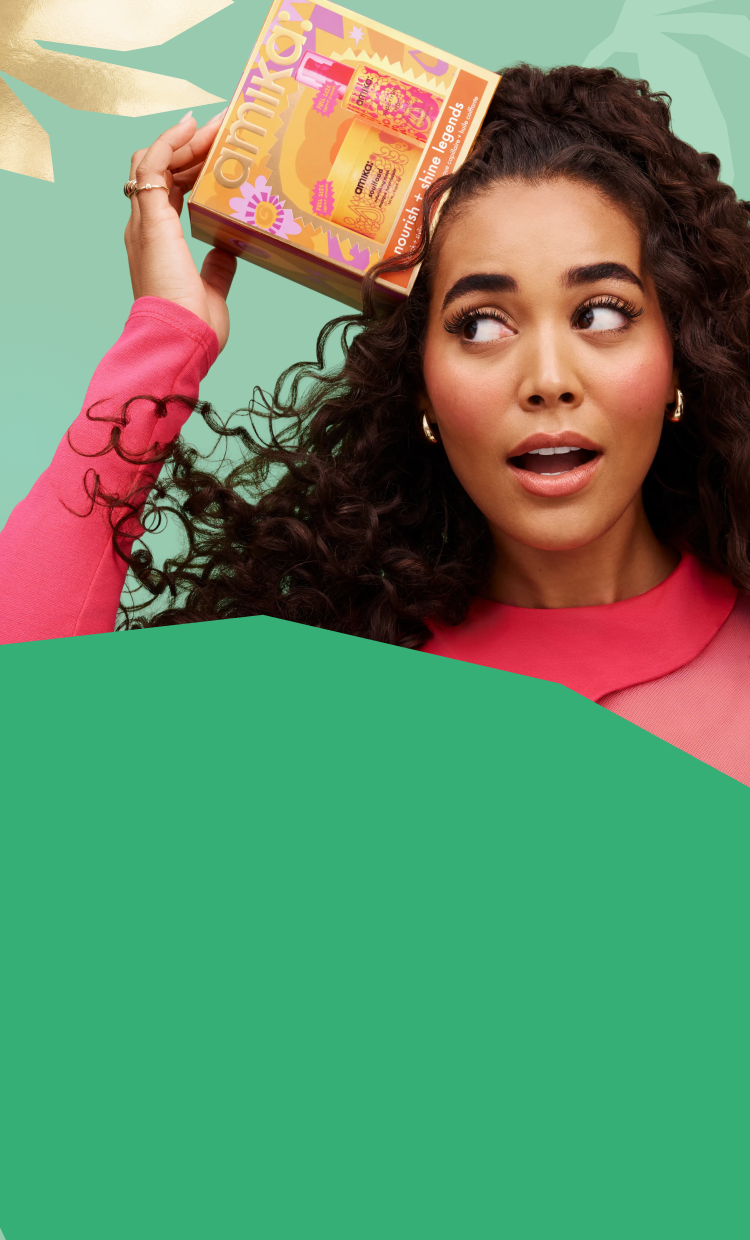 a model with long, brown, curly hair smiles while holding amika nourish + shine legends haircare gift set box. the background is green and there is a darker green shape in the foreground