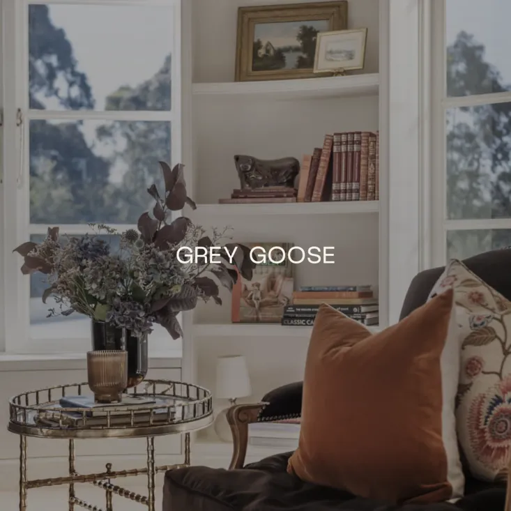 The Grey Goose wordmark in white over a sunlit sitting room with a bay window, a brass side table and a vase of dried flowers