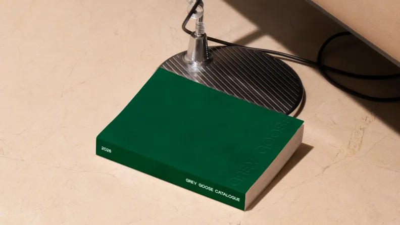 A green 2026 Grey Goose catalogue on a marble floor beside a lamp base