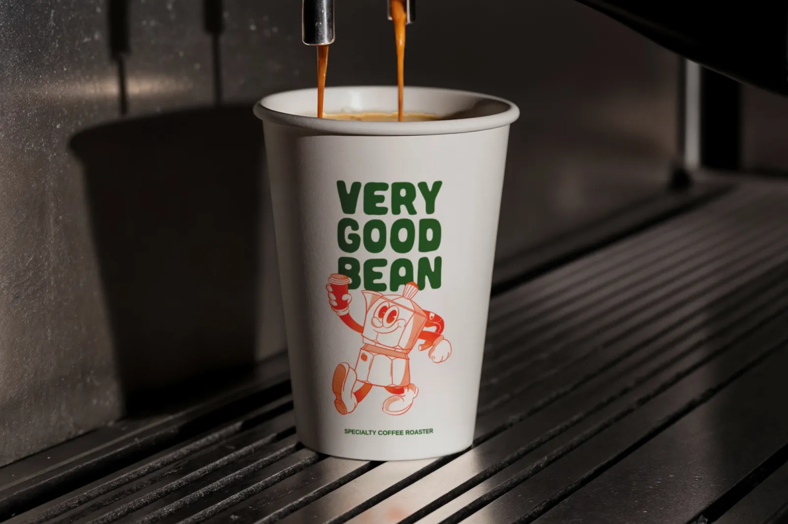 A Very Good Bean takeaway cup under an espresso machine, two streams of coffee pouring in, the green wordmark and orange moka pot character on the cup
