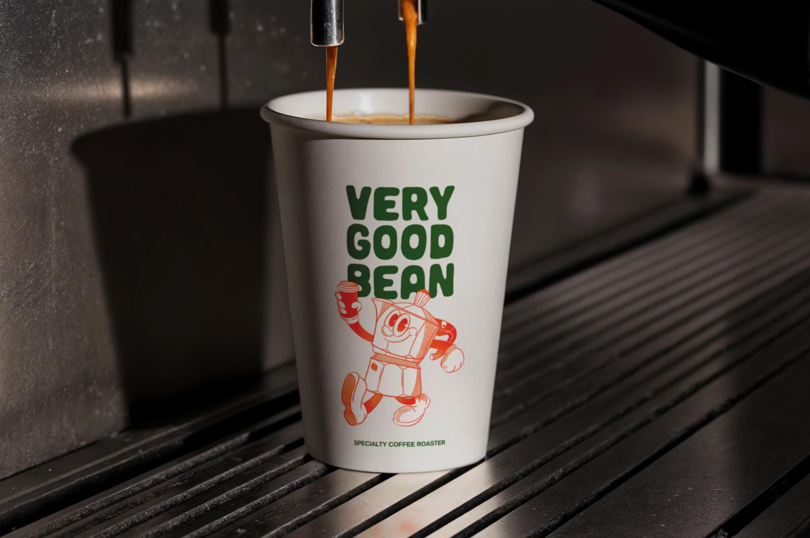 A Very Good Bean takeaway cup under an espresso machine, two streams of coffee pouring in, the green wordmark and orange moka pot character on the cup