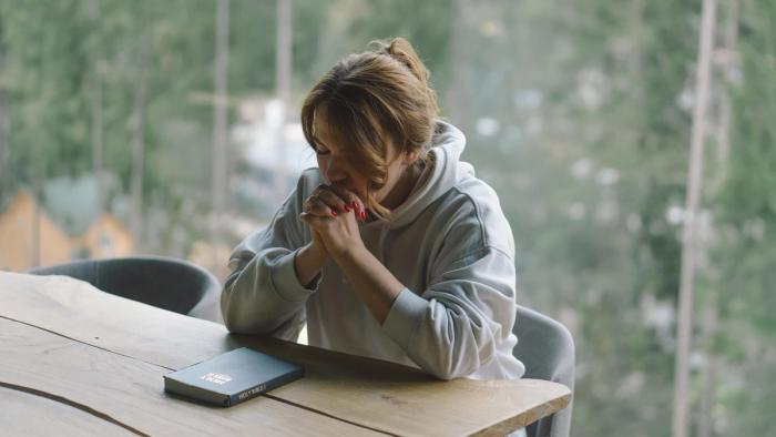 A woman praying with a Bible on the table in front of her.