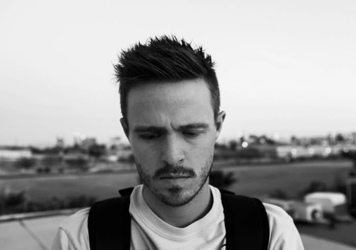 CC-  grayscale portrait of a down-cast or concerned, dark-haired young man with facial hair, wearing a white t-shirt and black vest against a nondescript outdoor background 