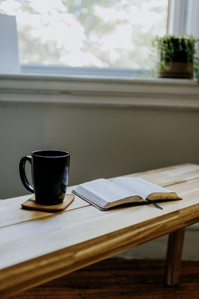 CC- a blonde-wood bench with a black mug of coffee on a leather coaster next to an open bible, under a window with a small potted succulent on the sill