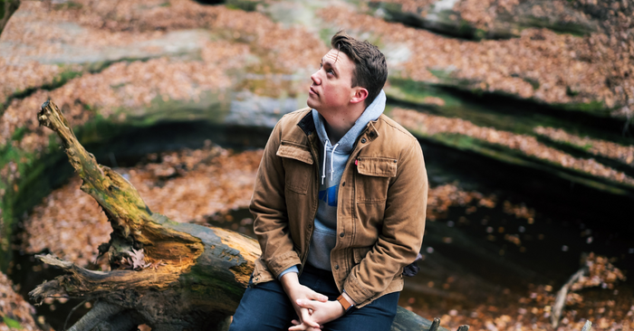 CC-  young man wearing an open tan canvas jacket over a gray hooded sweatshirt is seated on a moss-covered fallen tree, surrounded by moss-covered rock formation and many brown fallen leaves, looks up to his right