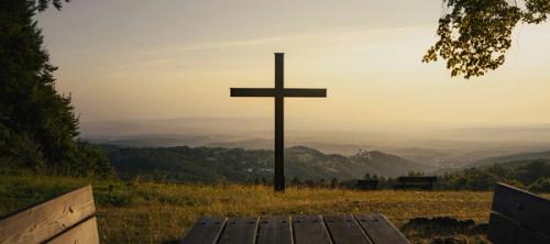 A cross in silhouette on a grassy hillside overlooking a valley.