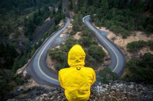 CC-  someone in a hooded yellow rain jacket sits on a high viewpoint overlooking a hairpin curve in a two-lane road cutting through a tree covered landscape