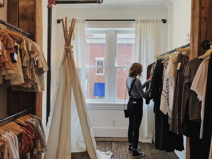 CC- two rows of closet rods of full of children's clothes hang along the left wall in a boutique clothing shop, a white-cloth teepee at the back wall, and to the right, a young woman in front of a sheer white curtain-flanked window, looks through women's clothes