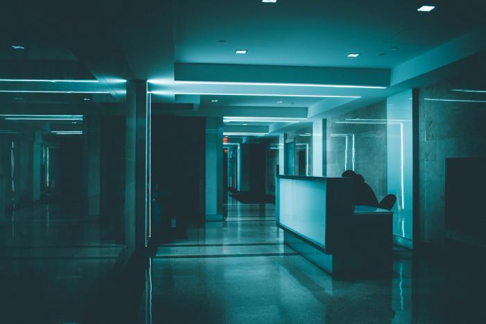 CC-  inside a dark building with lots of hard square lines and cold blue lighting, there is a lone dark-clad figure hunched behind a receptionist's desk