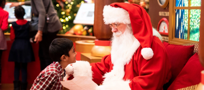 CC-  in a department store decorated for Christmas a young boy in a red paid shirt looks with happy intensity into the spectacled eyes of a seated older gentleman with a full white beard and mustache dressed as a santa