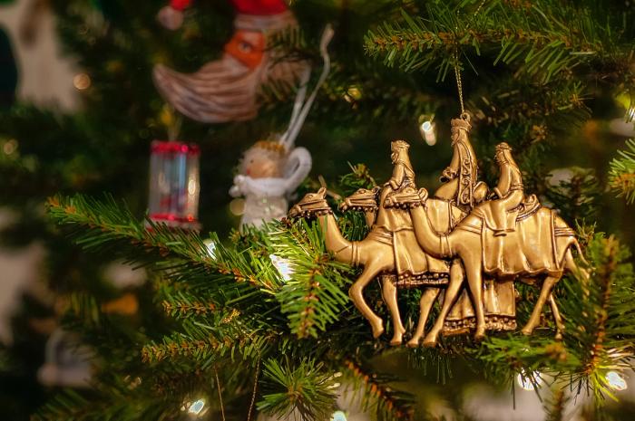 CC-  shallow focus on a golden ornament of a traditional rendering of three wise men on camels hanging from a branch of a Christmas tree lit with warm-white lights and various other blurry ornaments hang in the background