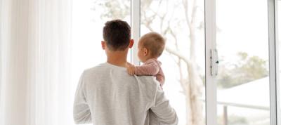 Back of man in white longsleeve holding blonde baby in red striped shirt in white room with windows