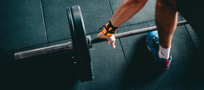 CC-  photo of the left end of a barbell set up on a gym floor with two plates held secure with a spring collar. The bar is being gripped by a man's hand wearing yellow and black wrist straps with a snatch grip