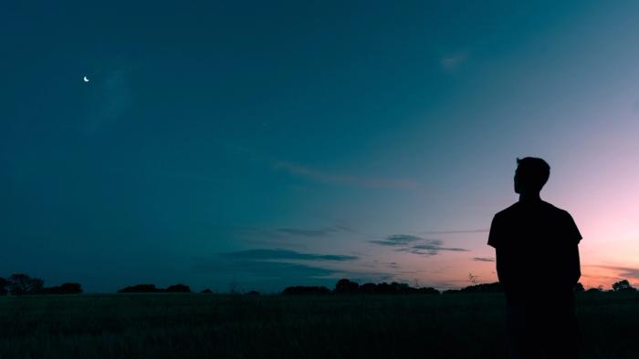 CC- a dark landscape stretches across the horizon at dusk with a male silhouette, backlit by a pink splash of sunset fading into a deep blue sky, looking up to the left at a crescent moon.