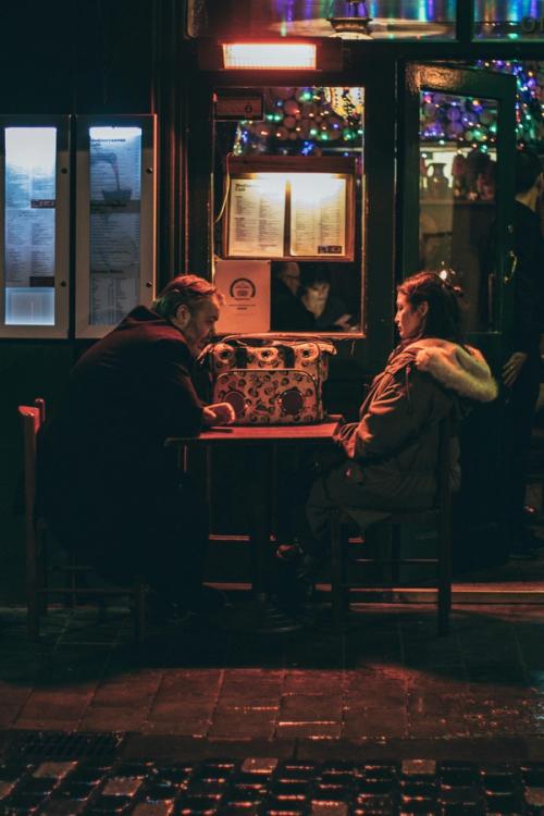 CC- An older couple sits at a table outside of a restaurant at night, engaged in what looks to be a serious conversation.