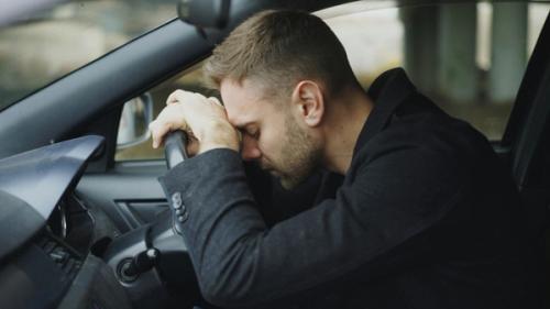 A young man sitting in his car looking depressed and resting his hands and head on the steering wheel.
