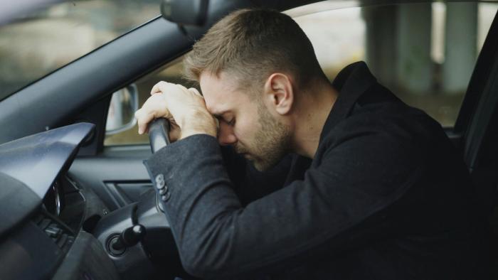 A young man sitting in his car looking depressed and resting his hands and head on the steering wheel.