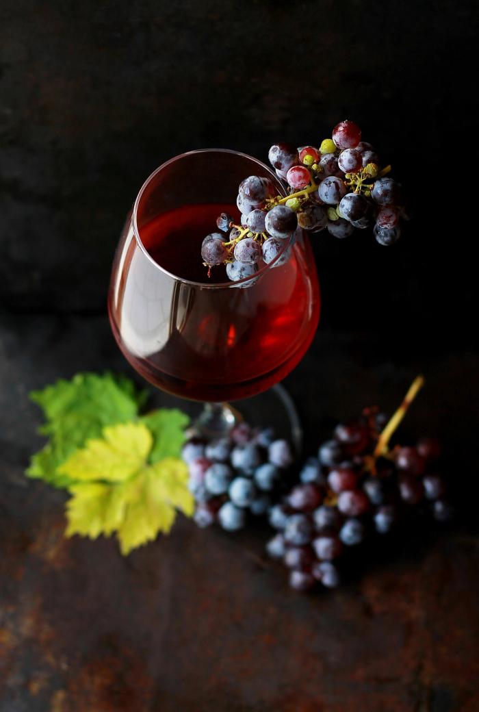 CC- a still life photograph of a wine glass filled with red wine and grapes immersed in the wine. There are grapes on the dark wood table too, as well as some fall foliage.