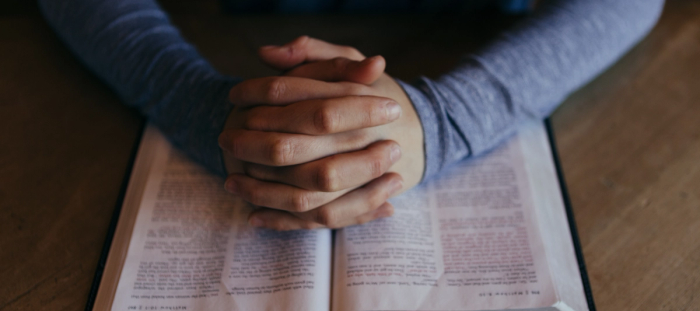 CC- hands folded resting on an open Bible on a light colored wood table top