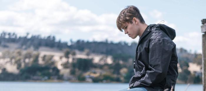 Young man in rain coat looking down sitting at the lake