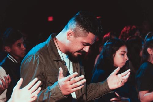 CC- A man stands in a dimly lit church service with his head down, eyes closed, and hands raised in prayer. He's surrounded by people in similar poses.