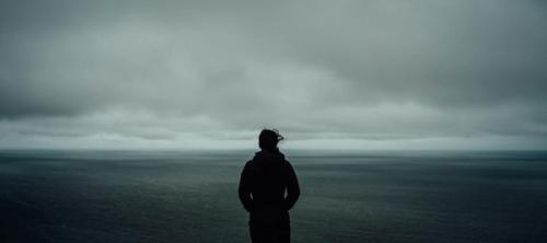 The silhouette of a person standing in a dreary, dark landscape with the wind blowing.