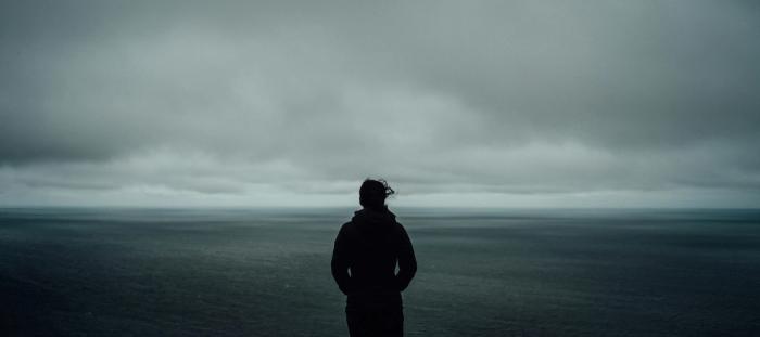 The silhouette of a person standing in a dreary, dark landscape with the wind blowing.