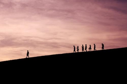 CC- A group of seven silhouetted people in the far distance follow behind a man walking ahead of them down a sloped road under a twilit sky.