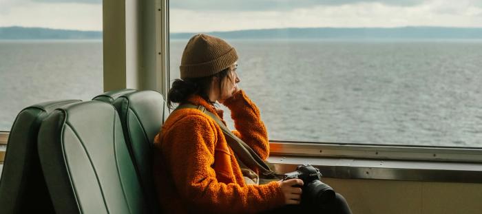 A woman by herself on a ferry looking out the window on an overcast day.