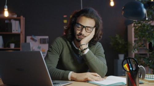A man falling asleep at his desk with his head resting on one hand.