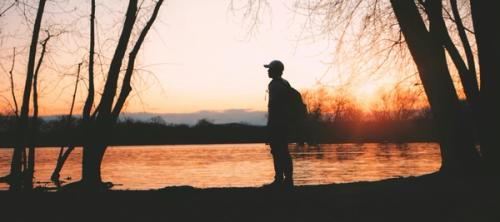 A man standing thoughtfully by a lake lit by a setting sun.