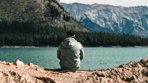 A man sitting on a rocky shore viewing a stunning lake with mountains in the distance.