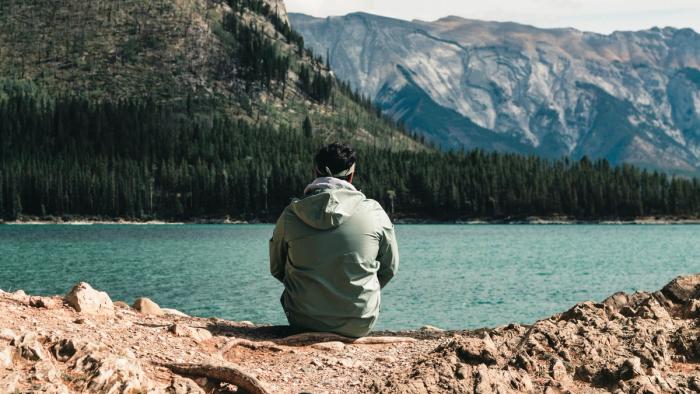 A man sitting on a rocky shore viewing a stunning lake with mountains in the distance.