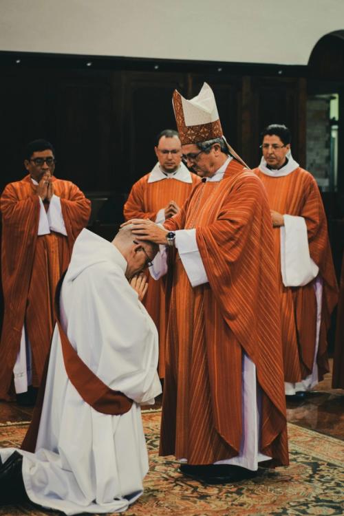 CC- A bishop blesses a man in a robe on his knees while other surrounding clergy watch.