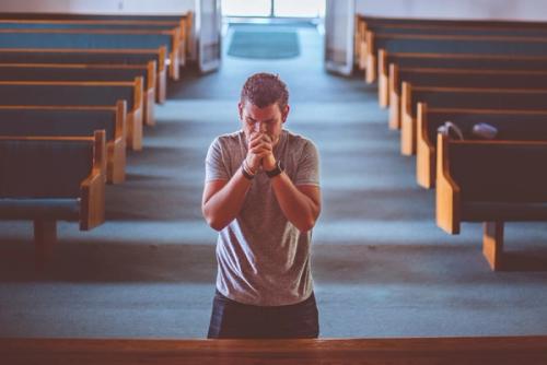 CC- A man stands at the front of a church with his hands clasped in prayer in front of his mouth.