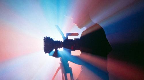 A man operating a professional video camera with blue and red light shining from behind.