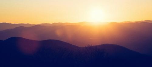 A bright sunrise in the distance over dark rows of hills.