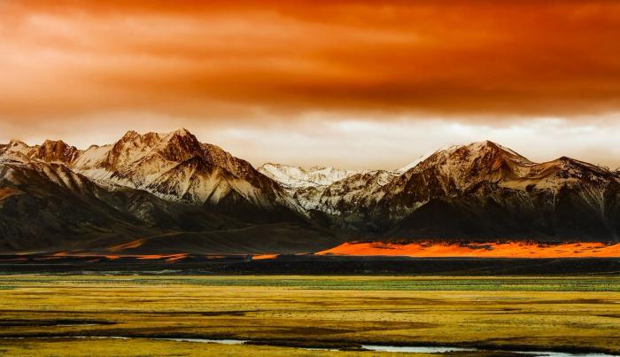 CC- panorama of a snow-dusted mountain range rising above a verdant valley in bold saturated colors into brilliant orange clouds