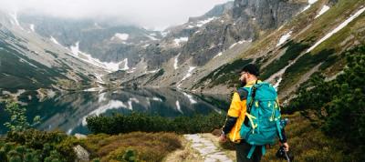 Man hiking in yellow shirt, black hat and teal back pack in the green valley beneath snowy mountains