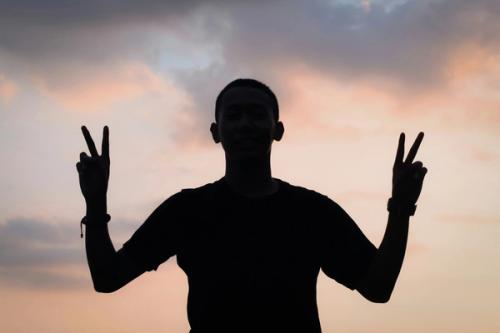 CC- A silhouetted man holds up two peace signs against a twilit sky.