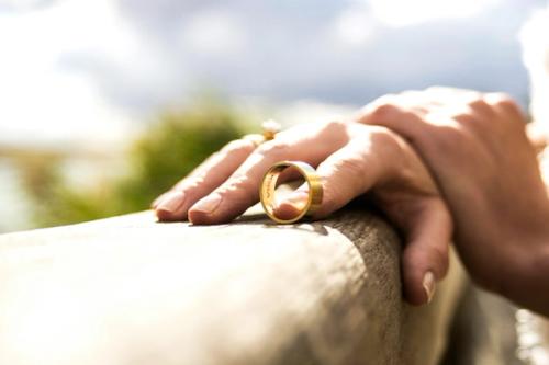 CC- A close up shot of a woman's hand supporting a man's wedding band on a wooden railing with the tip of her index finger.