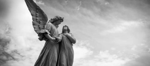 CC-  a monochromatic photo looking upward at a statue of a woman in a flowing robe holding a cross being supported by a winged being against a cloudy sky