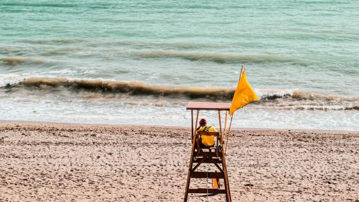 A lifeguard sitting in his tower at the ocean shore with a yellow flag flying.