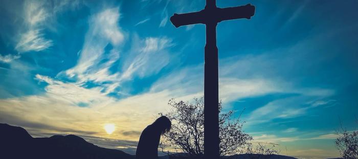 CC- silhouette of a cross and a person kneeling at its base backlit by a saturated blue sky and wispy white clouds