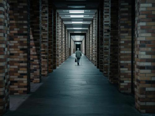 CC- An older man in the distance walks away from the camera in a cavernous stone hallway with pillars o