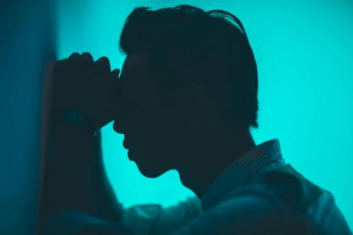 CC- A young man leans against a wall with his hands clasped and his eyes pressed into his hands. He's silhouetted against a bright window and ensconced in blue light.