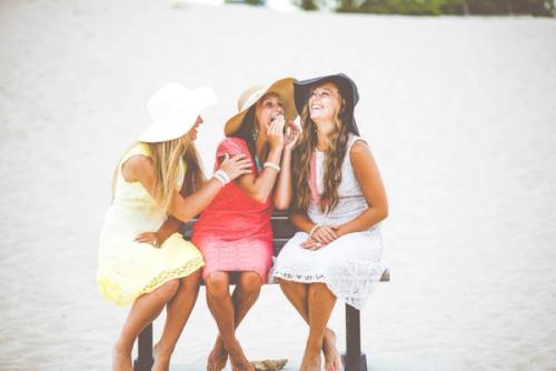 CC- Three ladies sit on a park bench in dresses and sun hats in front of a body of water laughing and whispering in each others' ears.