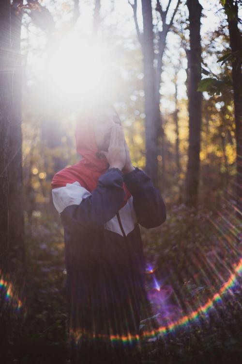 CC- A young man in a hoodie stands in a forest holding his hands together in prayer up to his mouth. The sun is shining through the trees and forming a rainbow lens flare effect around the man.