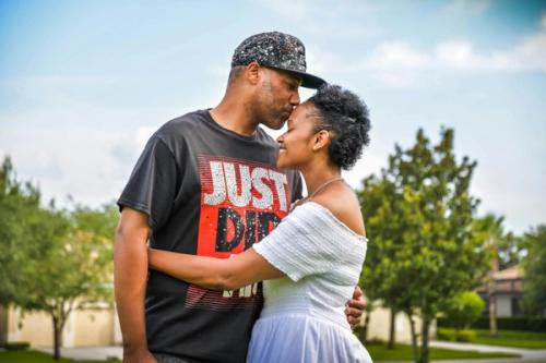 CC- A man kisses his smiling wife on the forehead as they stand in a suburb on a sunny day.